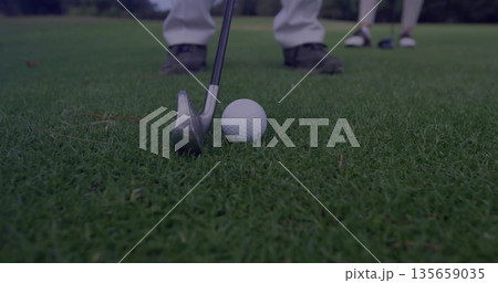Positioning steel golf iron behind white golf ball on putting green, with golf shoes over grass Positioning steel golf iron behind white golf ball on putting green, with golf shoes over grass 135659035