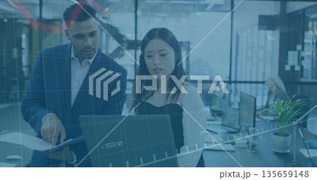 Collaborating colleagues reviewing laptop at open office, with printed report and monitors 135659148