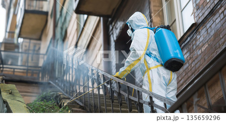 Worker in protective suit sprays pesticide on outdoor staircase of residential building, with greenery and brick wall visible in the background Worker in protective suit sprays pesticide on outdoor staircase of residential building, with greenery and brick wall visible in the background 135659296