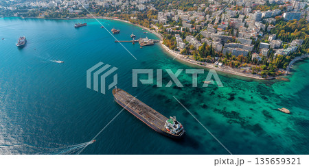 Aerial view of a coastal city with ships in the harbor and clear turquoise waters, surrounded by green hills and residential buildings along the shoreline Aerial view of a coastal city with ships in the harbor and clear turquoise waters, surrounded by green hills and residential buildings along the shoreline 135659321