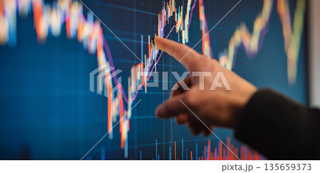 Hand of a person pointing at a financial chart displayed on a large screen, showcasing colorful candlestick patterns and data trends in a modern office environment 135659373