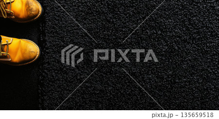 Yellow work boots with scuffed toes positioned on a textured black surface, showcasing the contrast between footwear and ground material in a close-up view 135659518