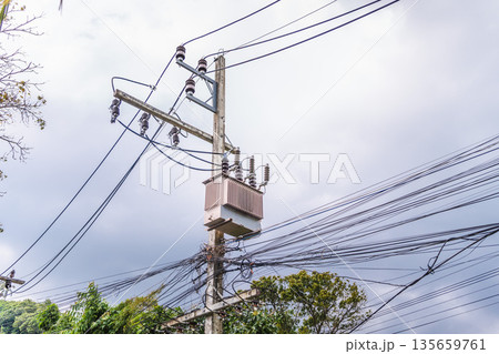 Electrical transformer and dense power lines on utility pole under cloudy sky, urban energy distribution infrastructure Electrical transformer and dense power lines on utility pole under cloudy sky, urban energy distribution infrastructure 135659761