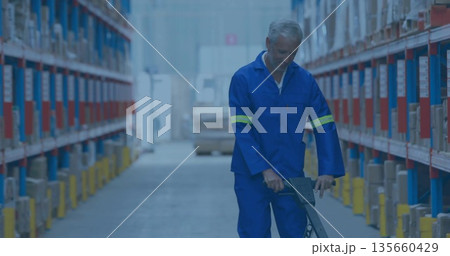 Warehouse worker in blue coveralls pushing pallet jack in storage aisle, with metal shelving racks Warehouse worker in blue coveralls pushing pallet jack in storage aisle, with metal shelving racks 135660429