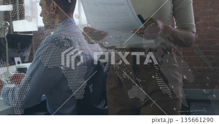 Collaborating analyst and manager reviewing reports on monitor at office, with world map overlay 135661290