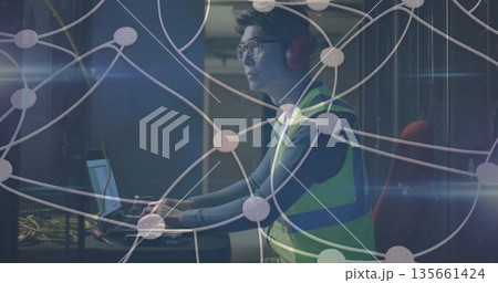 Technician wearing vest, headphones using laptop at data center, with server racks, network cables Technician wearing vest, headphones using laptop at data center, with server racks, network cables 135661424