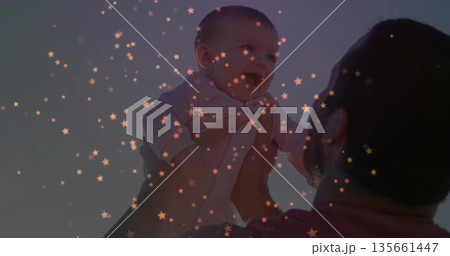 Father in casual shirt holding infant girl high above head in dusk sky, with star-shaped lights Father in casual shirt holding infant girl high above head in dusk sky, with star-shaped lights 135661447