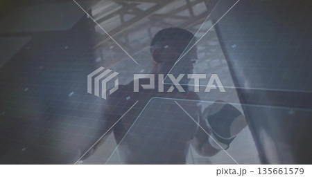 Male boxer wearing boxing gloves punching heavy bag in warehouse gym, with digital grid panels 135661579