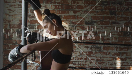 Leaning boxer wearing black sports bra and gloves, resting on ropes at gym, with brick wall 135661649