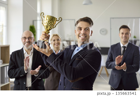 Happy businessman raising golden trophy with cheering colleagues in bright office 135663541