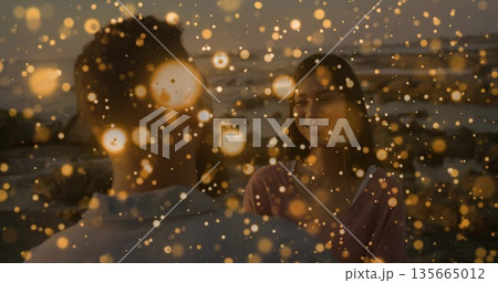 Smiling woman and man facing each other on shoreline at dusk, with golden fairy light bokeh 135665012