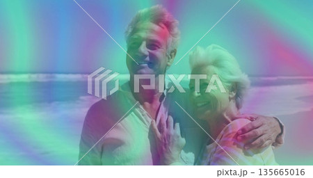 Smiling senior couple posing on sandy shoreline wearing light clothing, with waves rolling behind 135665016
