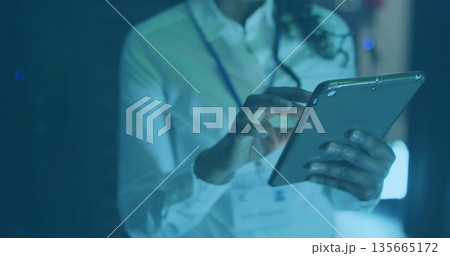 Mid adult woman monitoring equipment on tablet in control room, with ID badge and headset cable 135665172