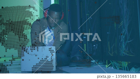 Typing technician wearing red headphones on silver laptop at data center with server racks, cables Typing technician wearing red headphones on silver laptop at data center with server racks, cables 135665183