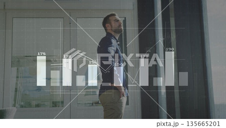 Standing man in modern office reviewing bar and line chart overlays on glass doors with blinds 135665201