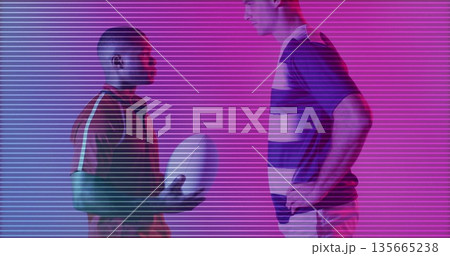 Posing rugby duo wearing sportswear holding white rugby ball at studio, with neon gradient lighting 135665238