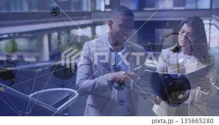 Pointing at network overlay, two suited professionals analyzing data inside atrium with tablet 135665280