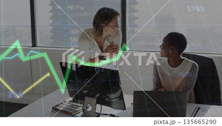 Discussing data, two business-casual women at desk in office, with monitor and financial chart 135665290
