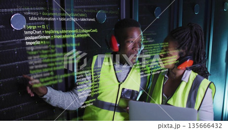 Technicians wearing safety vests pointing at server rack using laptop in data center, code overlay 135666432