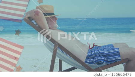 Lounging older man reclining on beach chair at shoreline, with striped swim trunks and straw fedora 135666475