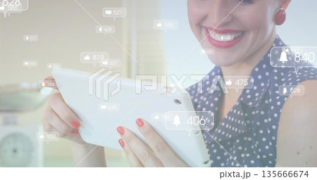 Smiling woman wearing navy blouse interacting with tablet in home kitchen, with notification icons 135666674