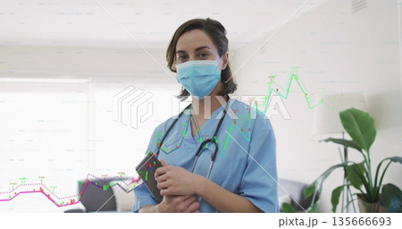 Standing worker wearing scrubs, mask and stethoscope holding tablet at clinic reception with plant Standing worker wearing scrubs, mask and stethoscope holding tablet at clinic reception with plant 135666693