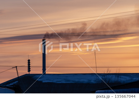 A chimney rising above the roof of a house is smoking. Smoke from the chimney against the backdrop of a winter sunset. A chimney rising above the roof of a house is smoking. Smoke from the chimney against the backdrop of a winter sunset. 135667044
