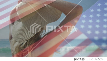 Gazing female beachgoer wearing straw sunhat and red bikini top at shoreline, with flag overlay 135667780
