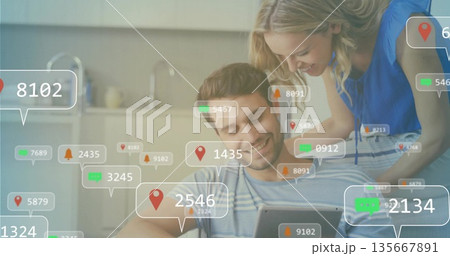 Tapping couple on sofa using tablet in open plan living room with floating notification icons 135667891