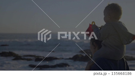 Standing mother carrying baby boy on shoulders at rocky shoreline, holding red bucket, copy space Standing mother carrying baby boy on shoulders at rocky shoreline, holding red bucket, copy space 135668186