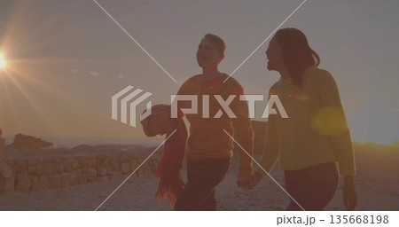 Walking couple holding hands along sandy shore at sunset, with rolled-up blanket and fringed scarf 135668198