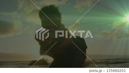 Walking man carrying woman along rocky shoreline at sunset, with ocean waves and rock formations 135668201