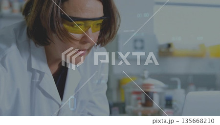 Female scientist wearing lab coat analyzing data at bench with glassware and laptop, copy space Female scientist wearing lab coat analyzing data at bench with glassware and laptop, copy space 135668210