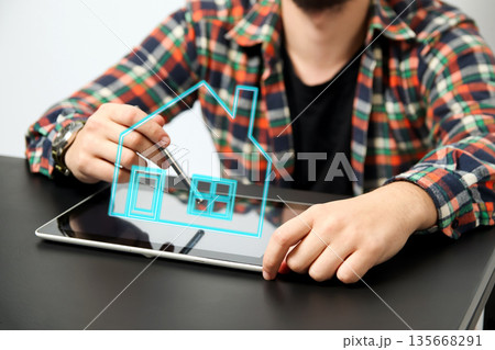 A man in a checkered shirt sits at a desk, using a stylus on a digital tablet. real estate search, smart home technology, or architectural planning A man in a checkered shirt sits at a desk, using a stylus on a digital tablet. real estate search, smart home technology, or architectural planning 135668291