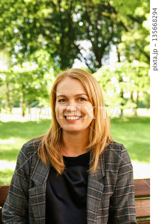 A portrait of a smiling middle-aged woman with blonde hair sitting on a wooden bench in a park. Vertical portrait woman 40s years A portrait of a smiling middle-aged woman with blonde hair sitting on a wooden bench in a park. Vertical portrait woman 40s years 135668294