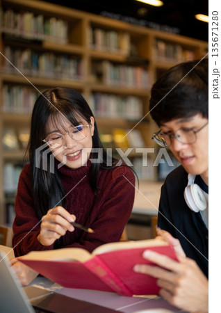 Glasses asian woman student pointing pencil in book man friend holding while sits at library's table 135671280
