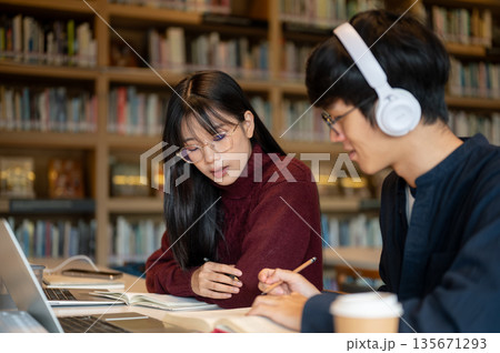 Glasses asian woman student helping a man friend doing assignment while sitting at library's table. 135671293