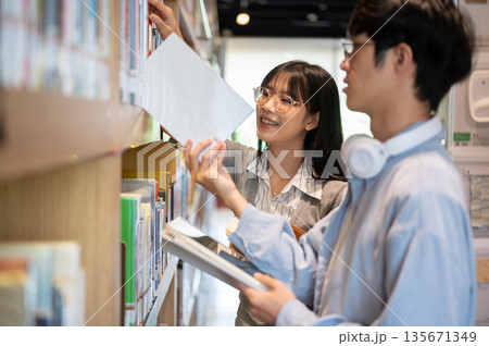 Glasses asian woman student and a man friend standing and looking through the library's bookshelf. Glasses asian woman student and a man friend standing and looking through the library's bookshelf. 135671349