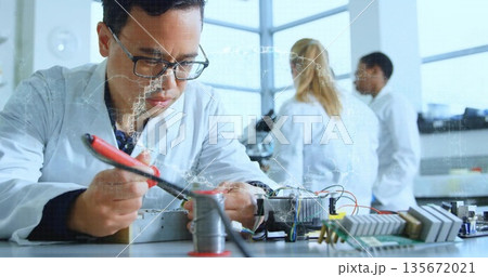 Soldering technician in lab coat using soldering iron leaning over lab bench with circuit boards 135672021