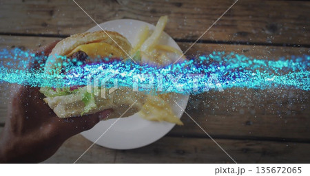 Lifting sesame-seed bun burger with adult hand over wooden tabletop, with ceramic plate of fries Lifting sesame-seed bun burger with adult hand over wooden tabletop, with ceramic plate of fries 135672065