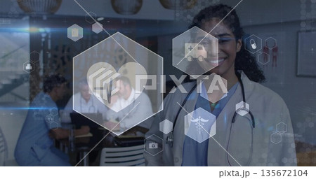 Smiling physician wearing white coat and stethoscope standing in clinic room, with medical icons 135672104