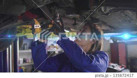 Working mechanic in blue coveralls using impact wrench under vehicle on hydraulic lift in workshop Working mechanic in blue coveralls using impact wrench under vehicle on hydraulic lift in workshop 135672417