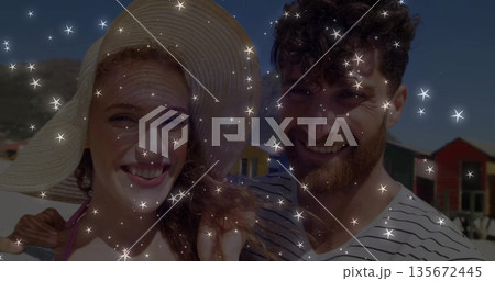 Smiling couple wearing straw hat on sandy shore, with colorful huts and sparkle overlay Smiling couple wearing straw hat on sandy shore, with colorful huts and sparkle overlay 135672445
