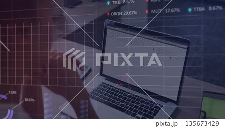 Mid-adult woman typing on silver laptop holding smartphone at desk, with floating chart overlays Mid-adult woman typing on silver laptop holding smartphone at desk, with floating chart overlays 135673429