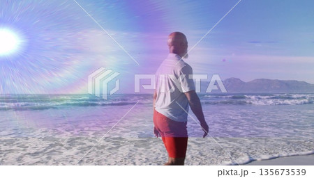 Gazing man wearing white T-shirt red trunks wading beach surf, with sun flare mountains, copy space Gazing man wearing white T-shirt red trunks wading beach surf, with sun flare mountains, copy space 135673539