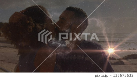 Kissing couple embracing on sandy beach at sunset, with sun flare, ocean waves and rock formations Kissing couple embracing on sandy beach at sunset, with sun flare, ocean waves and rock formations 135673632