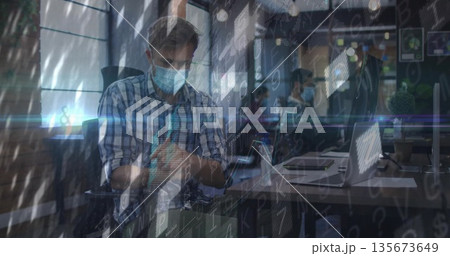 Man wearing shirt and mask applying hand sanitizer at office desk, with laptop and code overlay 135673649