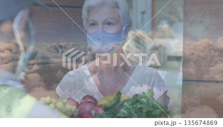 Senior masked woman accepting crate from vendor in safety vest at market stall, cloud overlay Senior masked woman accepting crate from vendor in safety vest at market stall, cloud overlay 135674869