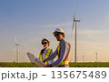 Two engineers in hard hats and safety vests review blueprints in  field with wind turbines at sunset on renewable energy projects 135675489