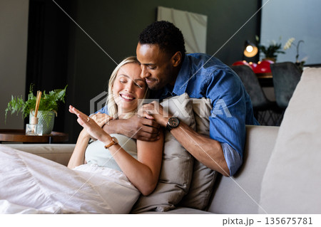 African American man hugging smiling woman on couch at home, enjoying togetherness 135675781